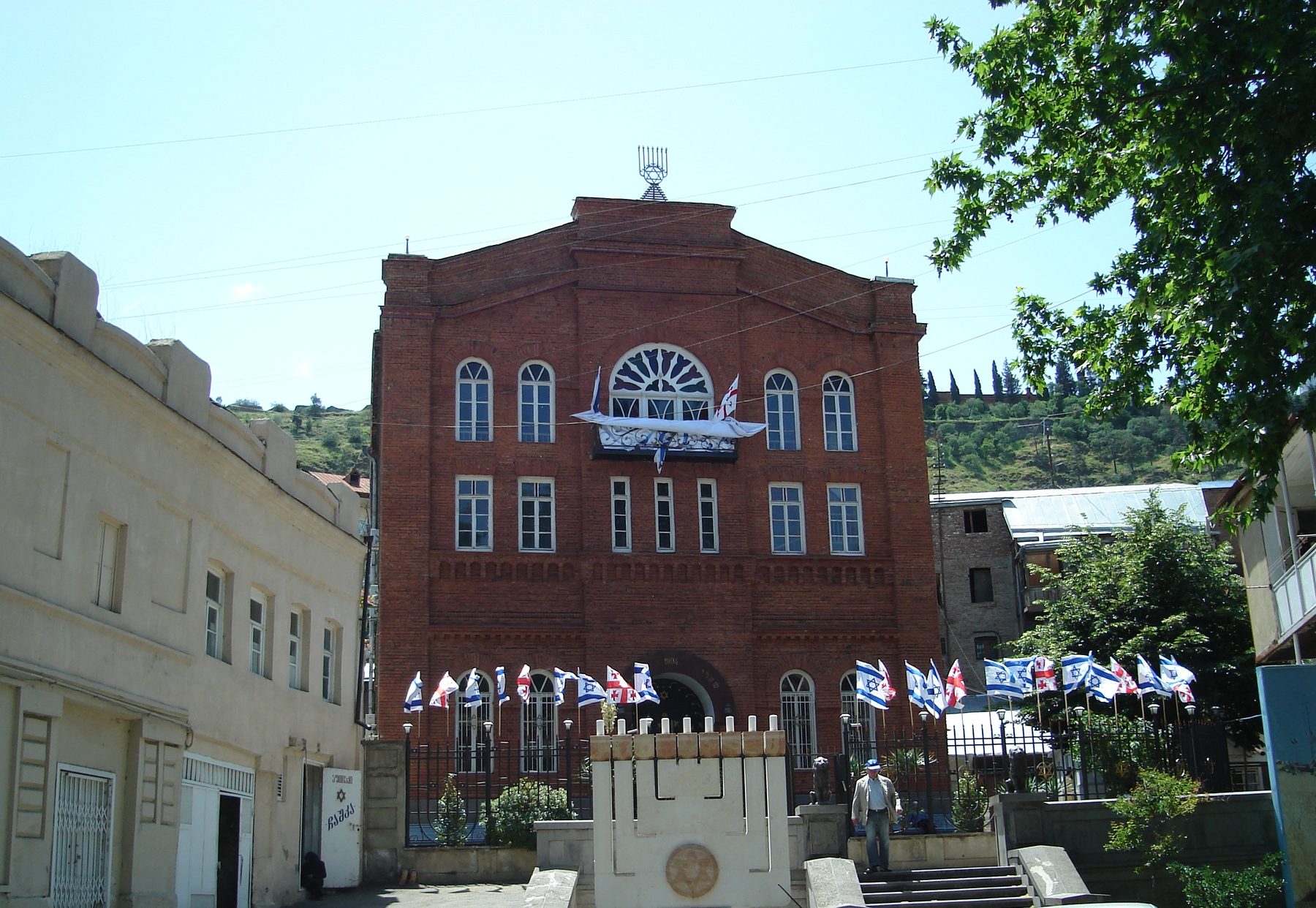 The red-brick Great Synagogue of Tbilisi, with a gabled facade topped by a menorah finial, Georgian and Israeli flags, and a Star-of-David monument in the forecourt